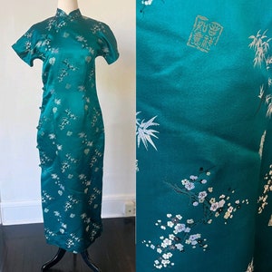 May include: Teal-colored, floor-length dress with short sleeves and a mandarin collar. The dress features a floral and bamboo pattern in white and silver. Buttons run down the side. The dress is displayed on a black mannequin.