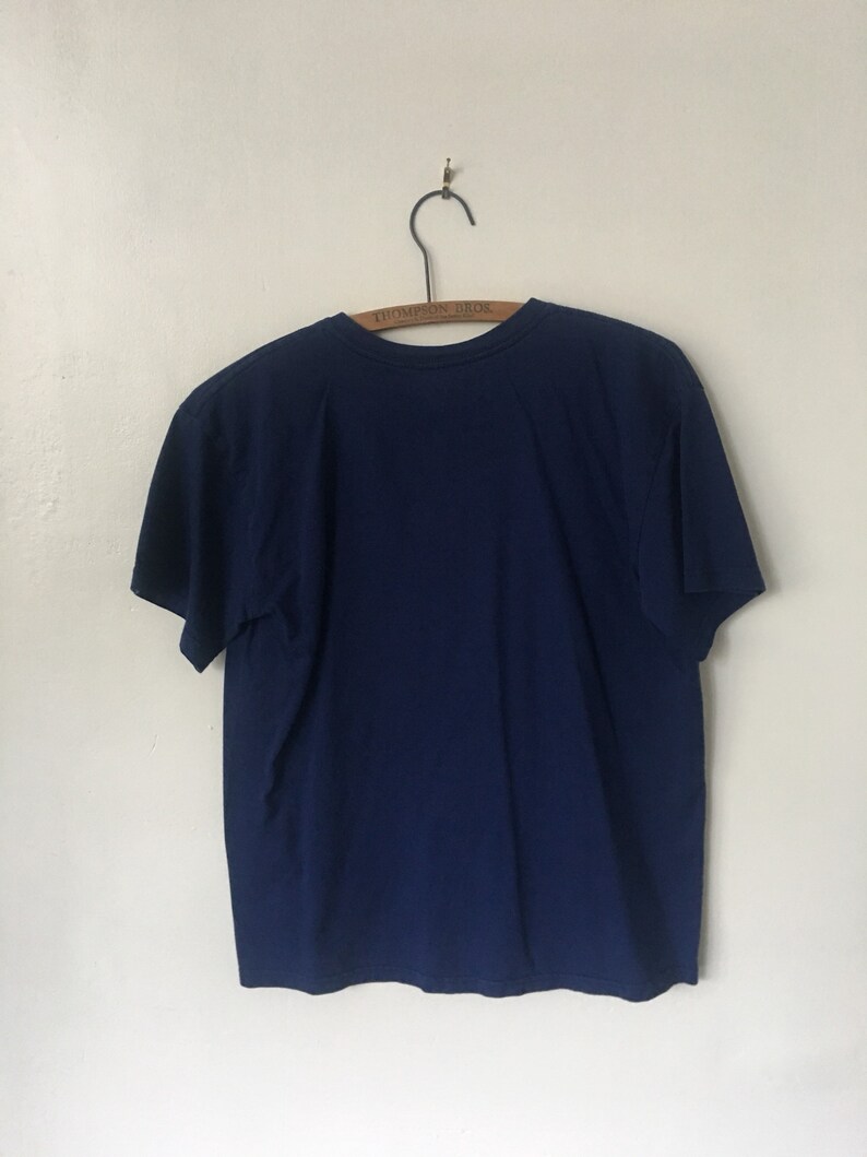 以下が含まれることがあります： 木製のハンガーにかけられた紺色のTシャツ。ハンガーには「Thompson Bros.」の文字が印刷されています。