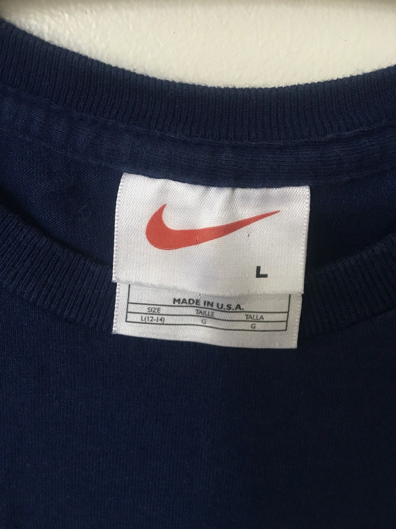 以下が含まれることがあります： 白地に「MADE IN U.S.A.」と赤いナイキのスウッシュロゴが書かれたタグが付いた紺色のTシャツ。タグには「SIZE L(12-14)」「TAILLE G」「TALLA G」の文字も書かれている。