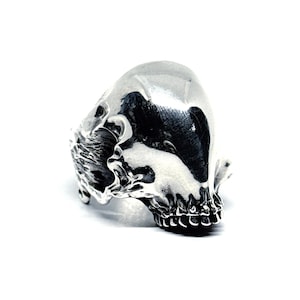 May include: A silver-colored ring in the shape of a skull. The skull ring has detailed features, including eye sockets, a nose cavity, and teeth. The ring is set against a white background, highlighting its metallic sheen and intricate design.