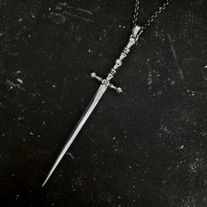 May include: A silver-toned pendant necklace featuring a miniature sword design. The sword has a detailed hilt and a long, pointed blade. The necklace hangs on a delicate chain, creating a gothic or fantasy-inspired accessory.
