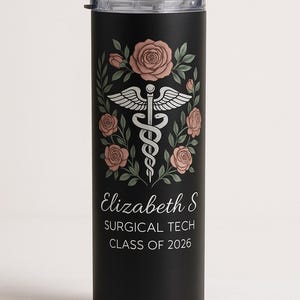 Op de afbeelding: Zwarte beker met een bloemenontwerp en het medische symbool. Het ontwerp bevat roze rozen en groene bladeren. De tekst luidt "Elizabeth S SURGICAL TECH CLASS OF 2026". Inclusief een zilveren rietje.