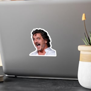 May include: A white sticker featuring a portrait of a man with dark curly hair, open mouth, and a surprised expression. The sticker is affixed to the lid of a laptop computer.