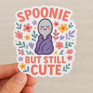 May include: A sticker with the text "SPOONIE BUT STILL CUTE" in coral. The sticker features a cartoon spoon wrapped in a purple blanket, surrounded by colorful flowers and leaves. The spoon has a smiling face.