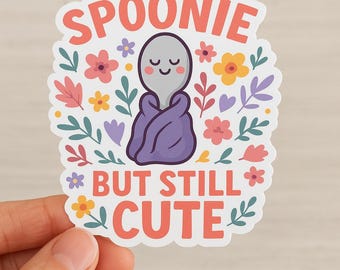 May include: A sticker with the text "SPOONIE BUT STILL CUTE" in coral. The sticker features a cartoon spoon wrapped in a purple blanket, surrounded by colorful flowers and leaves. The spoon has a smiling face.