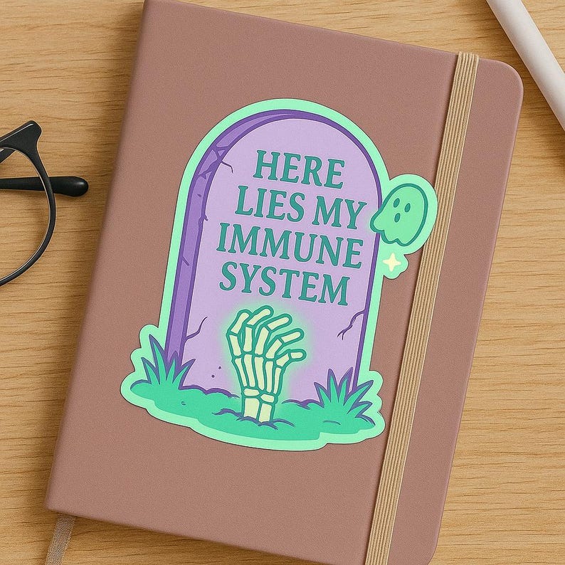 Funny Here Lies My Immune System sticker on mauve journal cover. Pastel glow spoonie vinyl decal with tombstone, ghost, and skeleton hand. Perfect sticker for planners, journals, and chronic illness humor gifts.