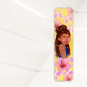 May include: A bookmark featuring a cartoon illustration of a woman with brown dreadlocks wearing pink glasses and a blue shirt. The background is yellow with pink flowers and butterflies.