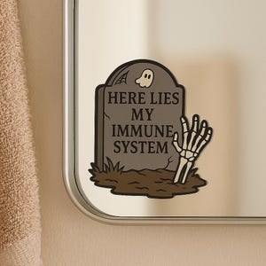 Dark graveyard Here Lies My Immune System sticker on bathroom mirror. Gothic tombstone design with skeleton hand and ghost, funny vinyl decal for spoonies and autoimmune warriors.