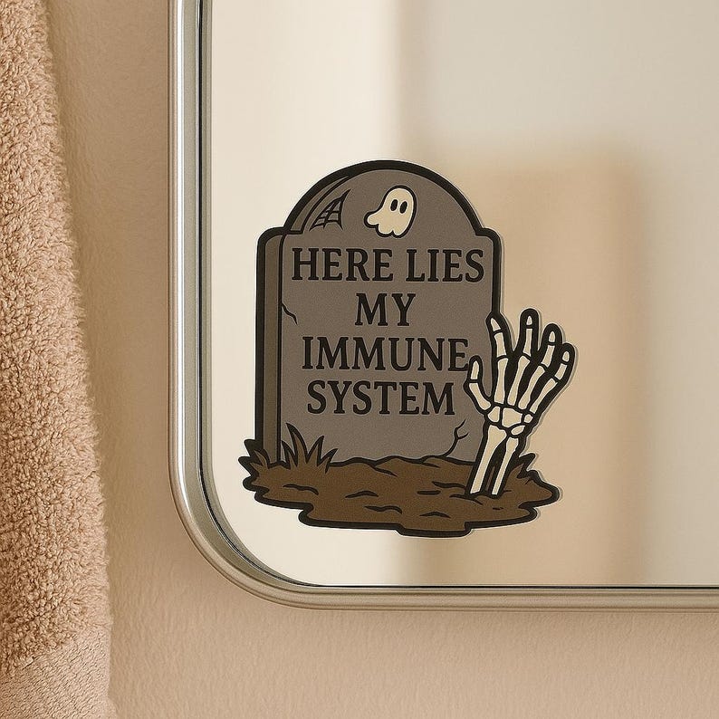 Dark graveyard Here Lies My Immune System sticker on bathroom mirror. Gothic tombstone design with skeleton hand and ghost, funny vinyl decal for spoonies and autoimmune warriors.