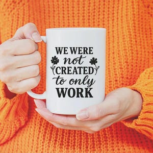 Könnte beinhalten: Weiße Keramik-Tasse mit dem schwarzen Text "WE WERE not CREATED to only WORK". Die Tasse wird von einer Person gehalten, die einen orangefarbenen Pullover trägt. Die Tasse hat einen weißen Henkel und ein Blumenmuster.