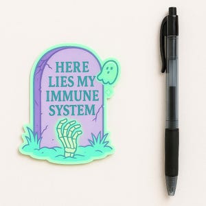 Funny pastel spoonie sticker reading Here Lies My Immune System shown next to black ballpoint pen for scale. 3x3 inch humorous vinyl decal for laptops, water bottles, and planners.