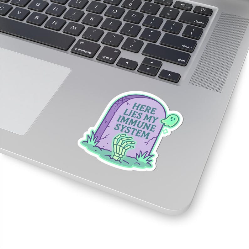Pastel Here Lies My Immune System sticker on silver laptop. Spoonie vinyl decal with lavender tombstone, mint ghost, and skeleton hand. Funny chronic illness sticker for laptops, planners, and water bottles.