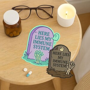 Pastel and dark Here Lies My Immune System stickers flat lay on nightstand with pill bottle, glasses, candle, and medications. Funny spoonie vinyl decals for chronic illness community, relatable gift.