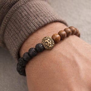May include: A beaded bracelet featuring a gold-colored lion head charm, black lava rock beads, and wooden beads. The bracelet is worn on a wrist, showcasing the contrast of materials and textures. A stylish accessory.