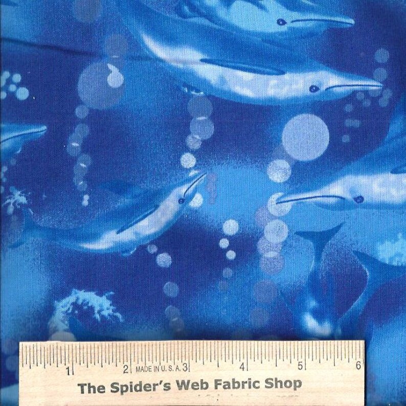 May include: Blue fabric with a repeating pattern of dolphins swimming in a blue ocean with white bubbles. The fabric is measured with a ruler that reads "The Spider's Web Fabric Shop".