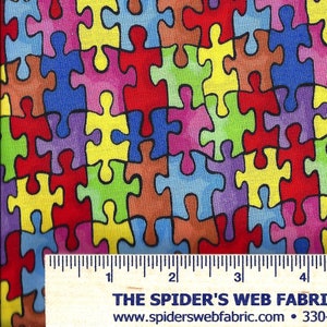 May include: A colorful fabric with a repeating pattern of puzzle pieces in various colors, including red, blue, yellow, green, and purple. The fabric is textured and has a slightly faded look. A ruler is visible at the bottom of the image, indicating the fabric is approximately 4 inches wide.