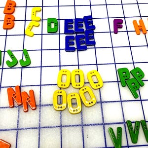 May include: Colorful alphabet letters scattered on a white grid. The letters are in various colors, including red, orange, yellow, green, blue, and purple. The letters are made of plastic and are used for educational purposes.