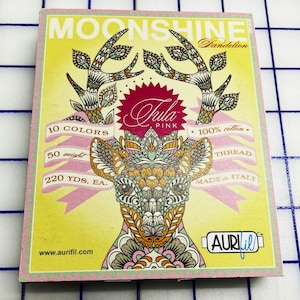 May include: A box of Aurifil Moonshine thread in the Tula Pink collection. The box features a decorative deer design with floral antlers and the text "10 Colors", "50 weight", and "220 YDS. EA." The thread is 100% cotton and made in Italy.