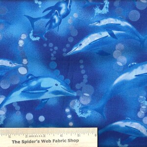 May include: Blue fabric with a repeating pattern of dolphins swimming in a blue ocean with white bubbles. The fabric has a ruler with the text "The Spider's Web Fabric Shop" printed on it.