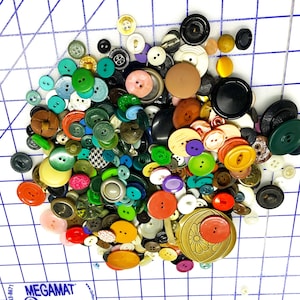 Może przedstawiać: Duża kolekcja kolorowych guzików w różnych kształtach, rozmiarach i materiałach. Guziki są mieszanką plastiku, metalu i tkaniny. Niektóre guziki mają dwa otwory, a inne cztery. Są też guziki z unikalnymi wzorami, np. kwiat.
