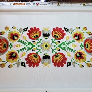 Puede incluir: Diseño bordado con un patrón simétrico de flores rojas, amarillas, verdes, blancas y negras. El diseño está centrado en un fondo blanco.