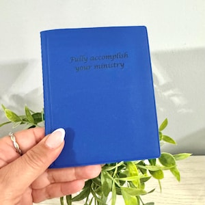 May include: A cobalt blue journal with the embossed text "Fully accomplish your ministry." The small book is held in a hand, with green foliage in the background. The journal is a solid color with a smooth texture.