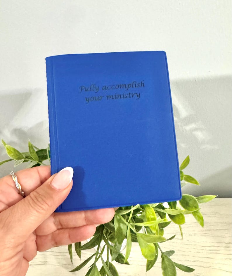 May include: A cobalt blue journal with the embossed text "Fully accomplish your ministry." The small book is held in a hand, with green foliage in the background. The journal is a solid color with a smooth texture.