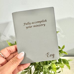 May include: A gray journal with the text "Fully accomplish your ministry" and the name "Evy" in a script font. The journal is held in front of a backdrop of green foliage.