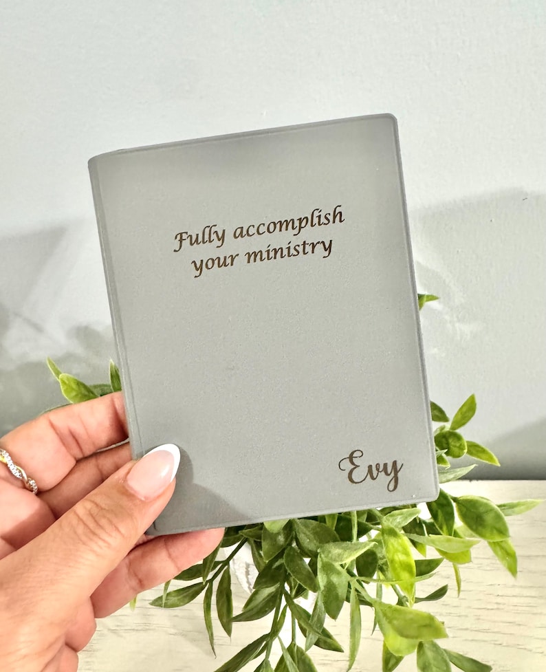 May include: A gray journal with the text "Fully accomplish your ministry" and the name "Evy" in a script font. The journal is held in front of a backdrop of green foliage.