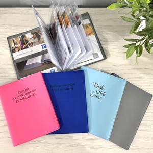 May include: A selection of small, colourful notebooks and a larger binder. The notebooks are in pink, blue, light blue, and grey, with black text. The binder is open, displaying pages. The text on the notebooks includes "Cumple completamente tu ministerio" and "Best Life Ever."