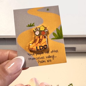 May include: A gold-colored enamel pin featuring a pair of orange work boots filled with colorful flowers. The pin is on a card with a yellow path design and the text "Your people will offer themselves willing! Psalm 110:3."