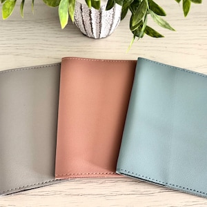 May include: Three leather passport holders in shades of gray, peach, and blue. The covers are smooth and stitched along the edges. The holders are arranged on a light wood-grain surface, with a decorative plant in the background.