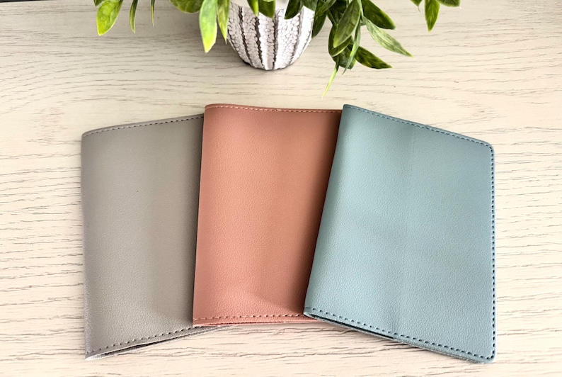 May include: Three leather passport holders in shades of gray, peach, and blue. The covers are smooth and stitched along the edges. The holders are arranged on a light wood-grain surface, with a decorative plant in the background.