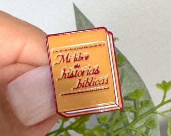 Puede incluir: Un pin de esmalte de color dorado con forma de libro y borde rojo. El libro tiene el texto "Mi libro de historias biblicas" escrito en la portada.