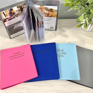 May include: A collection of colorful binders and clear plastic display cases. The binders are pink, blue, light blue, and gray, with text on the covers. The display cases hold printed cards with images and text.