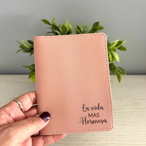 May include: A blush pink passport holder with the phrase "La vida mas Hermosa" in script. The cover is made of a smooth material and has a stitched border. The holder is held in a hand.