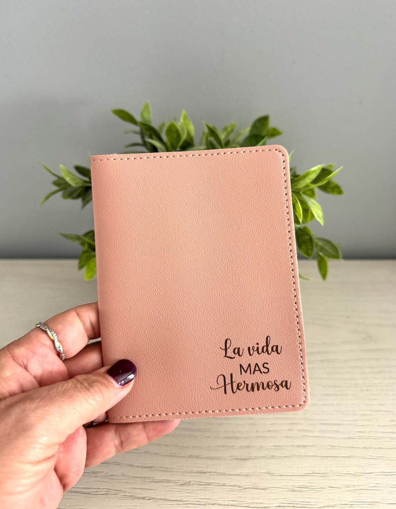 May include: A blush pink passport holder with the phrase "La vida mas Hermosa" in script. The cover is made of a smooth material and has a stitched border. The holder is held in a hand.