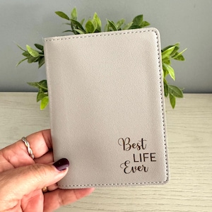 May include: A light gray passport holder with the text "Best Life Ever" in a script font. The holder is rectangular with stitched edges. The background includes green leaves.