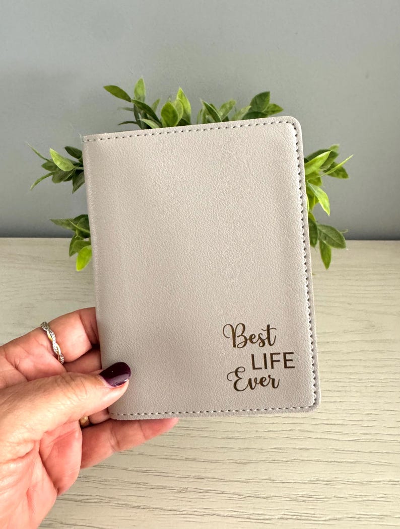 May include: A light gray passport holder with the text "Best Life Ever" in a script font. The holder is rectangular with stitched edges. The background includes green leaves.