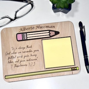 Könnte beinhalten: Schreibtisch-Organizer aus Holz mit Bleistift-Design, gelbem Haftnotizblock und Stift. Der Organizer trägt den Namen "Alberto Hussman" und einen eingravierten Bibelvers. Eine Brille befindet sich im Hintergrund.