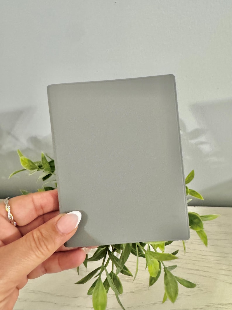 May include: A rectangular, gray, matte-finish object is held in front of green leafy sprigs. The object is smooth and appears to be a small, flat notepad or a decorative piece. The background is a light gray wall.