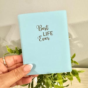 May include: A light blue notebook with the words "Best Life Ever" in elegant script. The notebook is held in a hand with a silver ring, surrounded by green foliage. The cover is a solid, soft blue color.