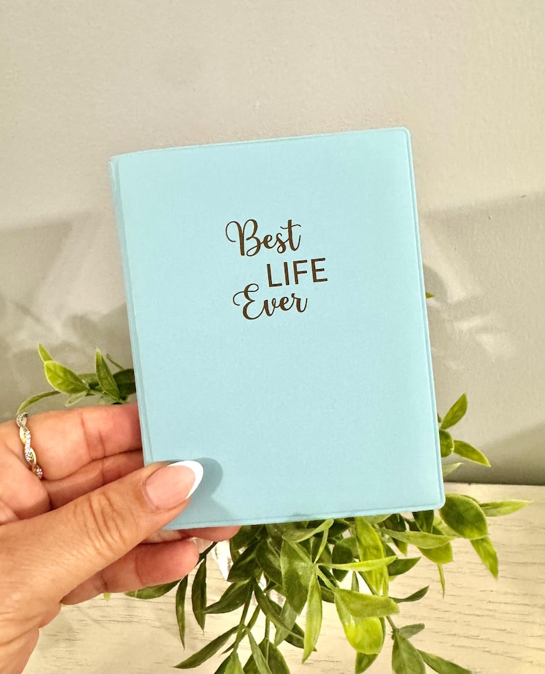 May include: A light blue notebook with the words "Best Life Ever" in elegant script. The notebook is held in a hand with a silver ring, surrounded by green foliage. The cover is a solid, soft blue color.