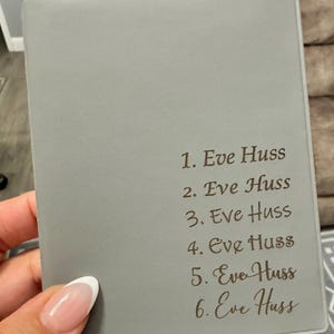 May include: A gray notebook with the name "Eve Huss" repeated six times in a brown script font. The notebook is held in a hand with a French manicure. The background is blurred.