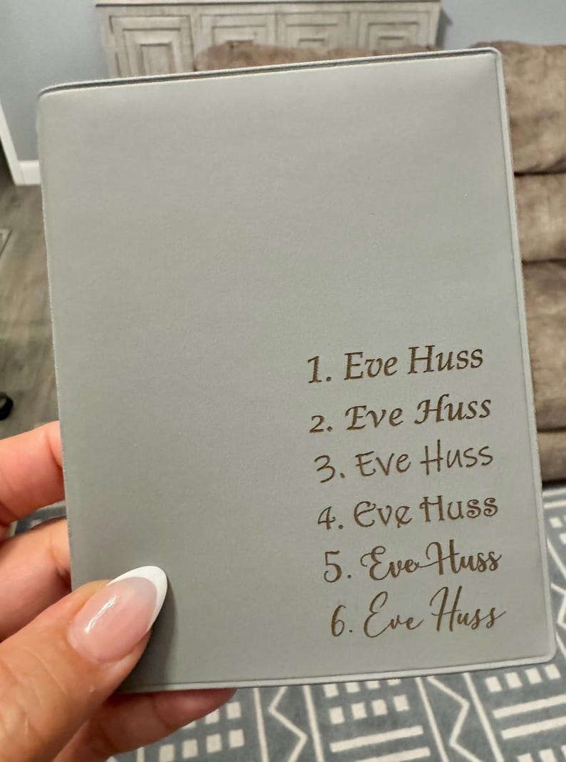 May include: A gray notebook with the name "Eve Huss" repeated six times in a brown script font. The notebook is held in a hand with a French manicure. The background is blurred.