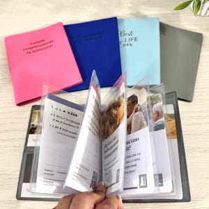 May include: A collection of small, colorful notebooks with text on the covers. The notebooks are pink, blue, and light blue. An open booklet with clear plastic sleeves is in the foreground, displaying printed pages with images and text. The text on the pink notebook reads "Cumple completamente tu ministerio."