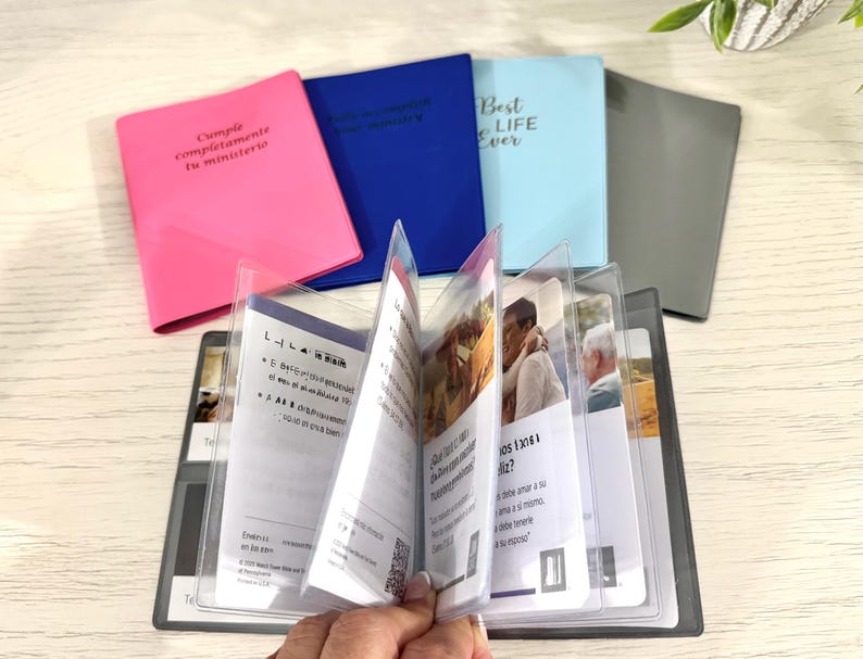 May include: A collection of small, colorful notebooks with text on the covers. The notebooks are pink, blue, and light blue. An open booklet with clear plastic sleeves is in the foreground, displaying printed pages with images and text. The text on the pink notebook reads "Cumple completamente tu ministerio."