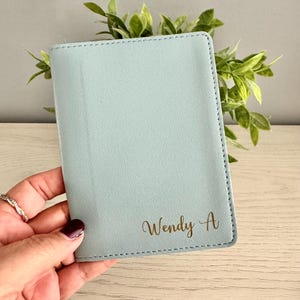 May include: A light blue passport holder with the name "Wendy A" in gold script. The holder is rectangular with stitched edges. Green foliage is visible in the background.