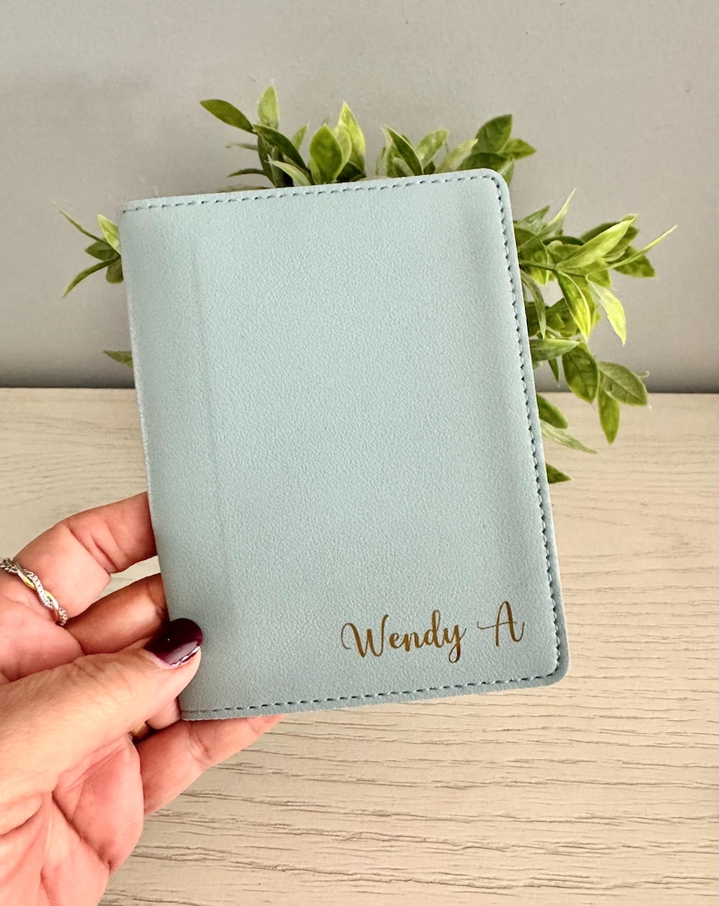 May include: A light blue passport holder with the name "Wendy A" in gold script. The holder is rectangular with stitched edges. Green foliage is visible in the background.