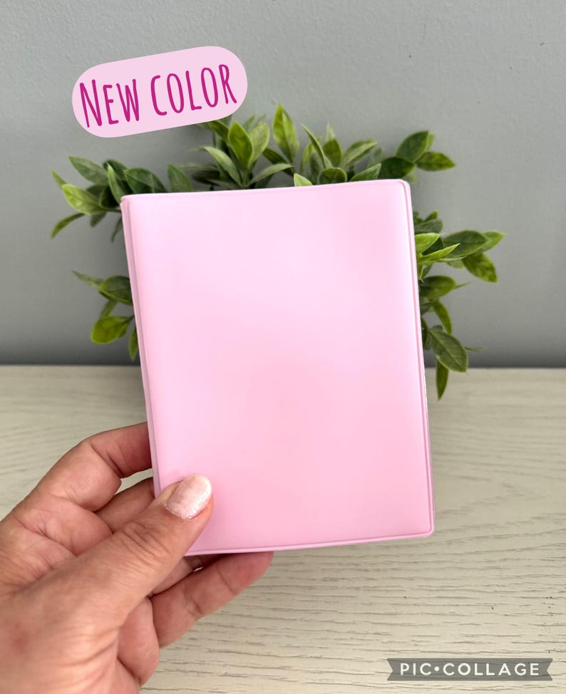 May include: A light pink rectangular object, possibly a wallet or small notebook, is held in a hand. The text "NEW COLOR" is in a pink oval above the object. Green leafy plants are in the background.